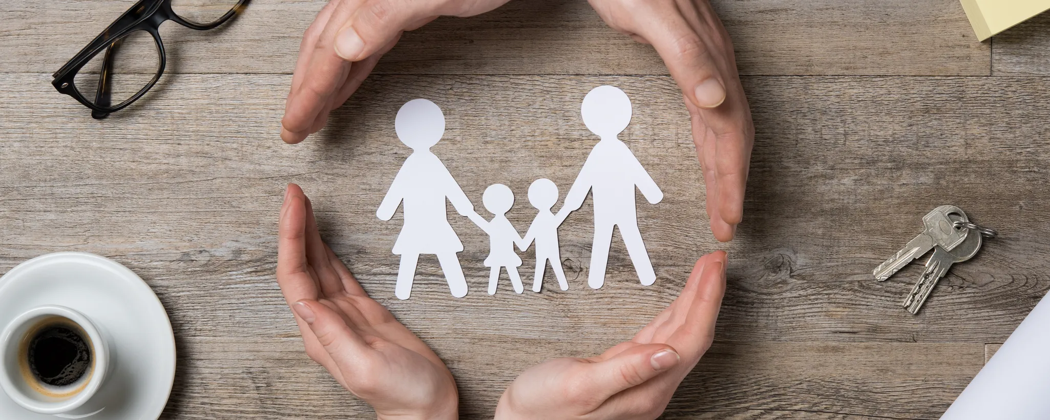 Zwei Hände formen einen schützenden Kreis um ausgeschnittene Papierfiguren einer Familie, symbolisierend den Sofortschutz. Auf dem Tisch liegen eine Brille, ein Kaffeetasse und Schlüssel.