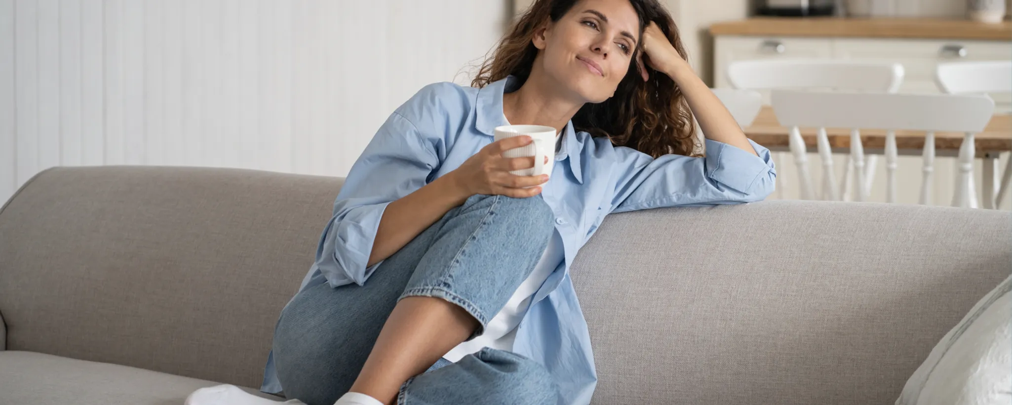 Eine Person sitzt entspannt auf einem grauen Sofa, trägt eine blaue Bluse und Jeans und hält eine Tasse in der Hand. Im Hintergrund ist ein Essbereich zu sehen, der an einen Versicherer erinnert, der für Sicherheit und Komfort steht.