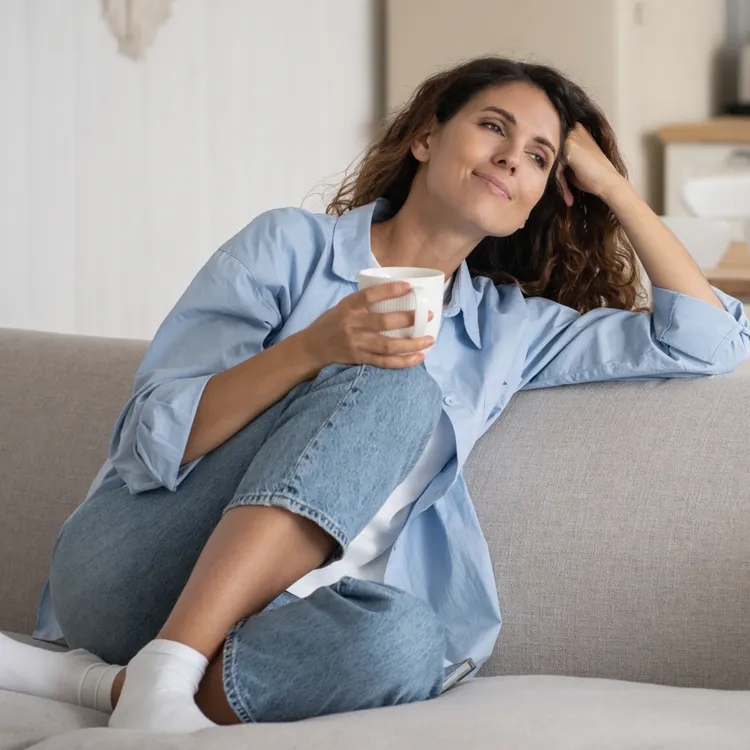 Eine Person sitzt entspannt auf einem grauen Sofa, trägt eine blaue Bluse und Jeans und hält eine Tasse in der Hand. Im Hintergrund ist ein Essbereich zu sehen, der an einen Versicherer erinnert, der für Sicherheit und Komfort steht.