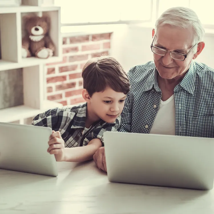 Ein älterer Mann und ein Junge sitzen gemeinsam am Tisch, beide vertieft in ihre Laptops. Im Hintergrund ist ein Regal mit einem Teddybär sichtbar. Sie diskutieren möglicherweise über die Rürup-Rente und Riester-Rente, um einen Überblick über die Altersvorsorge zu gewinnen.