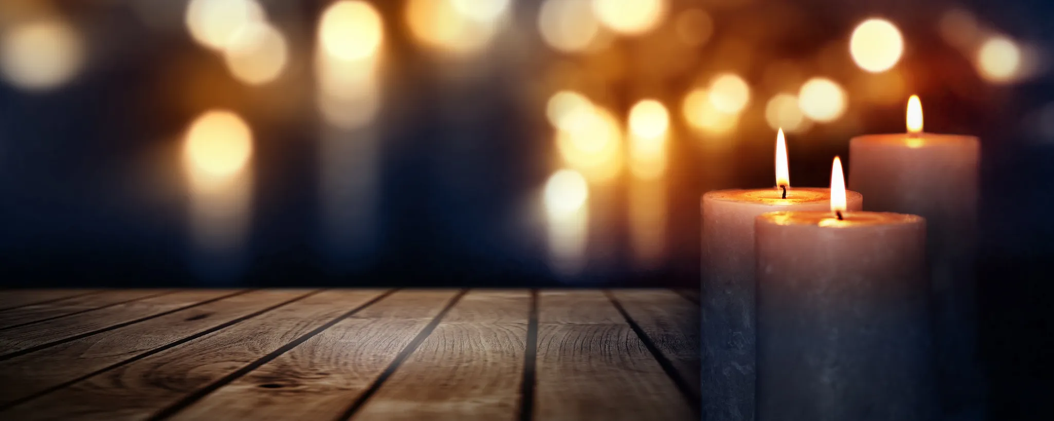 Drei brennende Kerzen auf einem Holztisch, die warmes Licht spenden. Sie symbolisieren oft Ruhe und Besinnung, insbesondere bei einem Todesfall. Im Hintergrund sind unscharfe Lichtpunkte sichtbar.