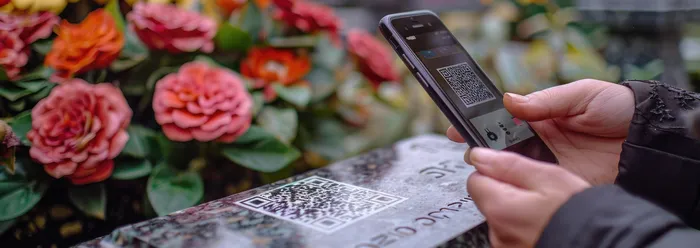 Eine Person hält ein Smartphone und scannt einen QR-Code auf einem Grabstein, während im Hintergrund blühende Rosen zu sehen sind. Der QR-Code ist mehrfach im Bild vorhanden.