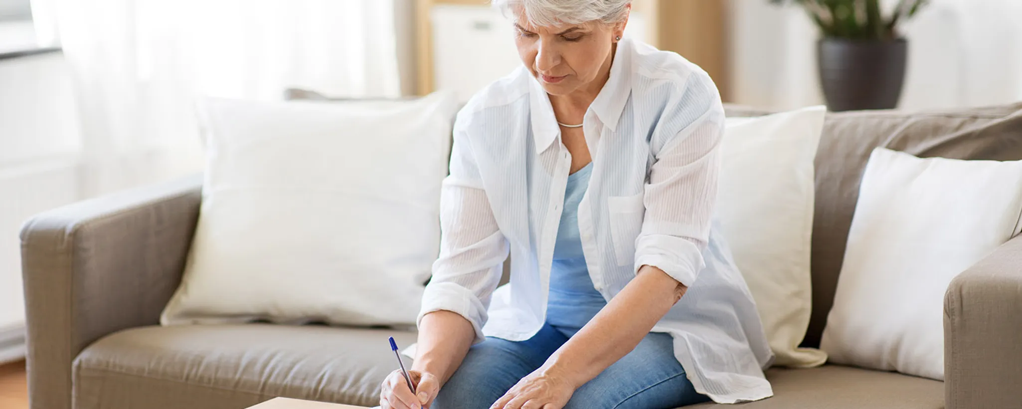 Eine ältere Frau sitzt auf einem beigen Sofa und macht Notizen auf einem Blatt Papier. Im Hintergrund sind helle Kissen sichtbar, die eine gemütliche Atmosphäre schaffen. Die Rentenlücke könnte ein Thema ihrer Notizen sein.