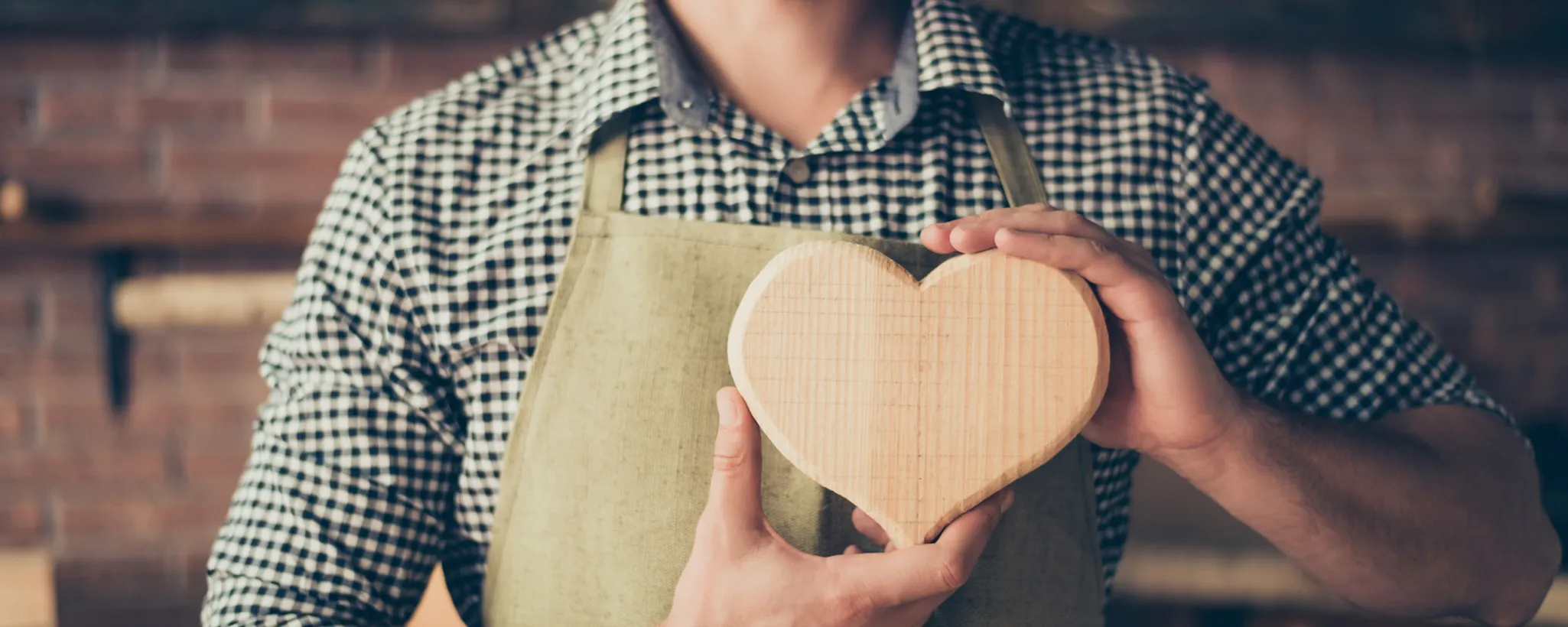 Ein Mann in kariertem Hemd und Schürze hält ein handgefertigtes Holzherz. Solche Kunstwerke sind bei Deutschen beliebt und symbolisieren oft Sicherheit und Geborgenheit, ähnlich wie Versicherungen.