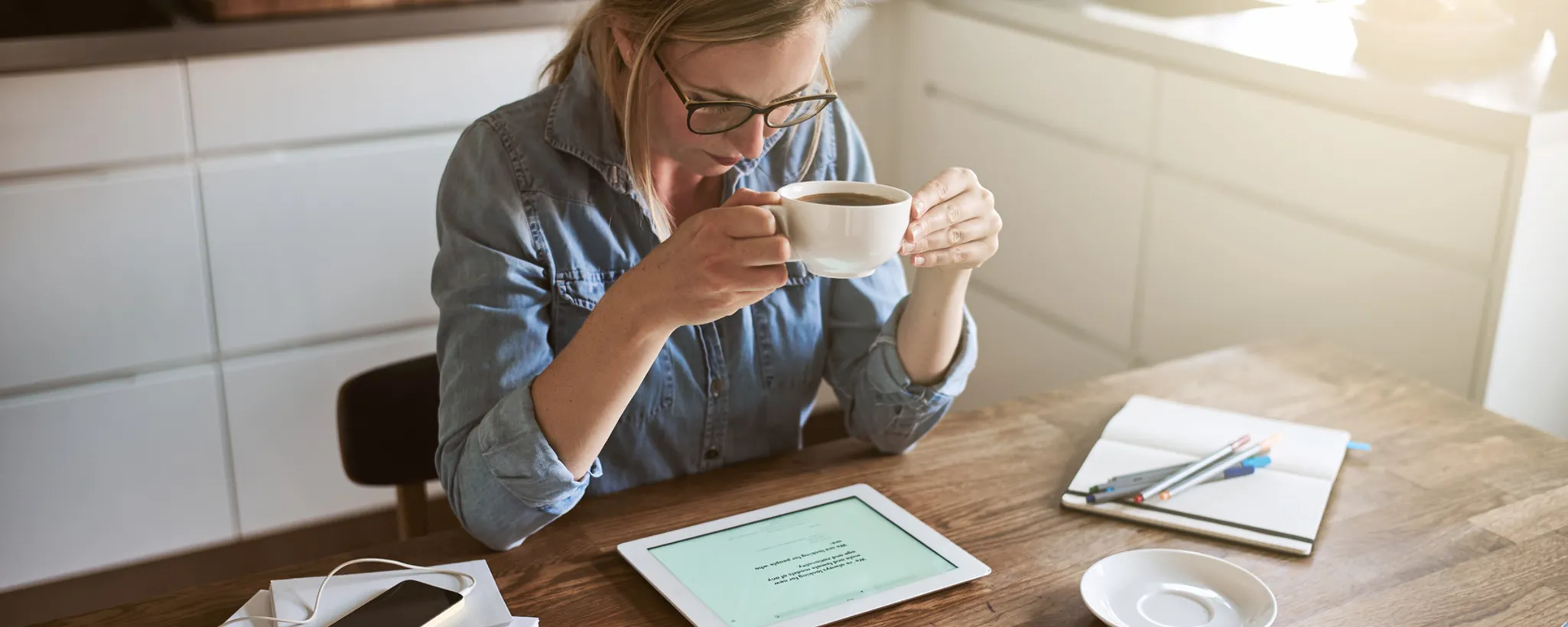 Eine Person sitzt an einem Holztisch, trinkt Kaffee und betrachtet ein Tablet, auf dem Informationen zu Versicherungen für Selbstständige angezeigt werden. Daneben liegen Notizbuch und Smartphone.