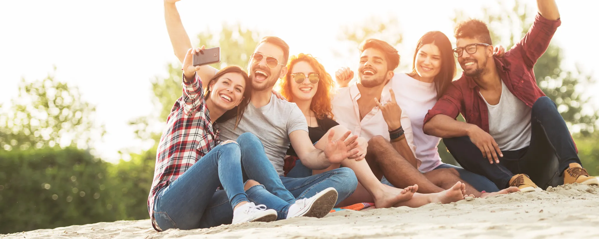 Eine Gruppe junger Studierender sitzt fröhlich im Sand und macht ein Selfie. Im Hintergrund sind Bäume zu sehen, die eine entspannte Atmosphäre schaffen. Versicherungen spielen hier keine Rolle.