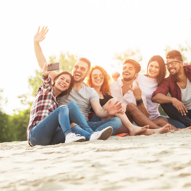 Eine Gruppe junger Studierender sitzt fröhlich im Sand und macht ein Selfie. Im Hintergrund sind Bäume zu sehen, die eine entspannte Atmosphäre schaffen. Versicherungen spielen hier keine Rolle.