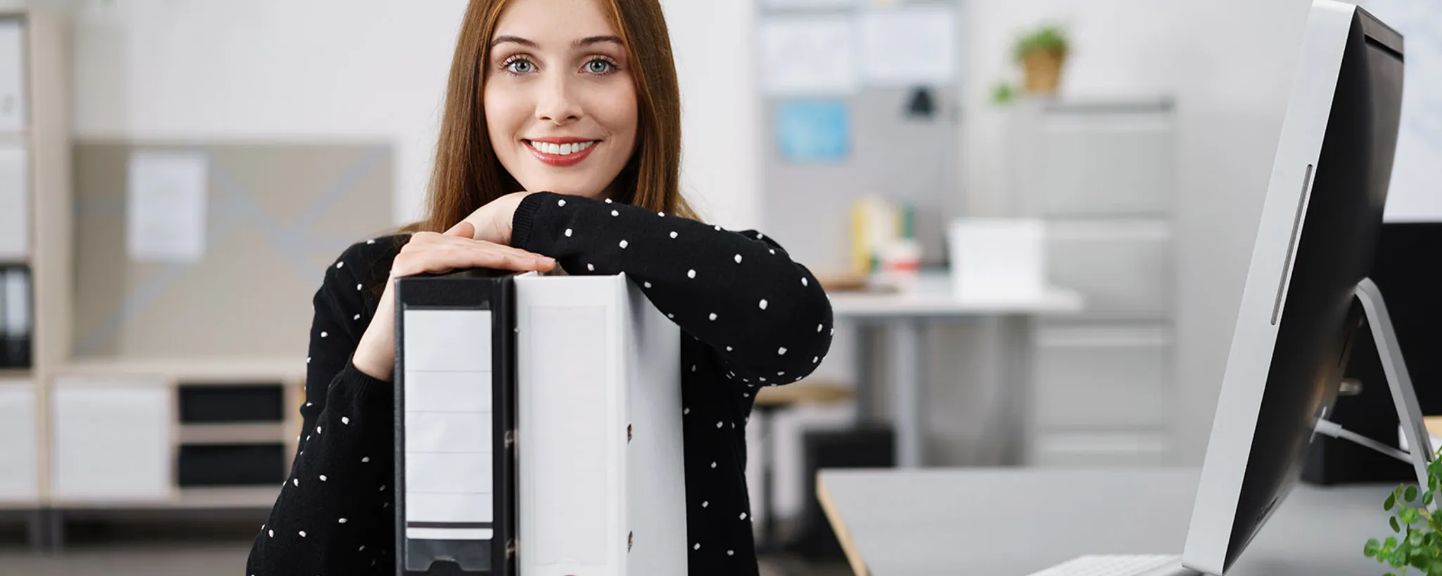 In einem modernen Büro hält eine Person zwei große Ordner in den Händen. Im Hintergrund sind ein Schreibtisch und eine Ablage mit Pflanzen und Büromaterialien zu sehen.