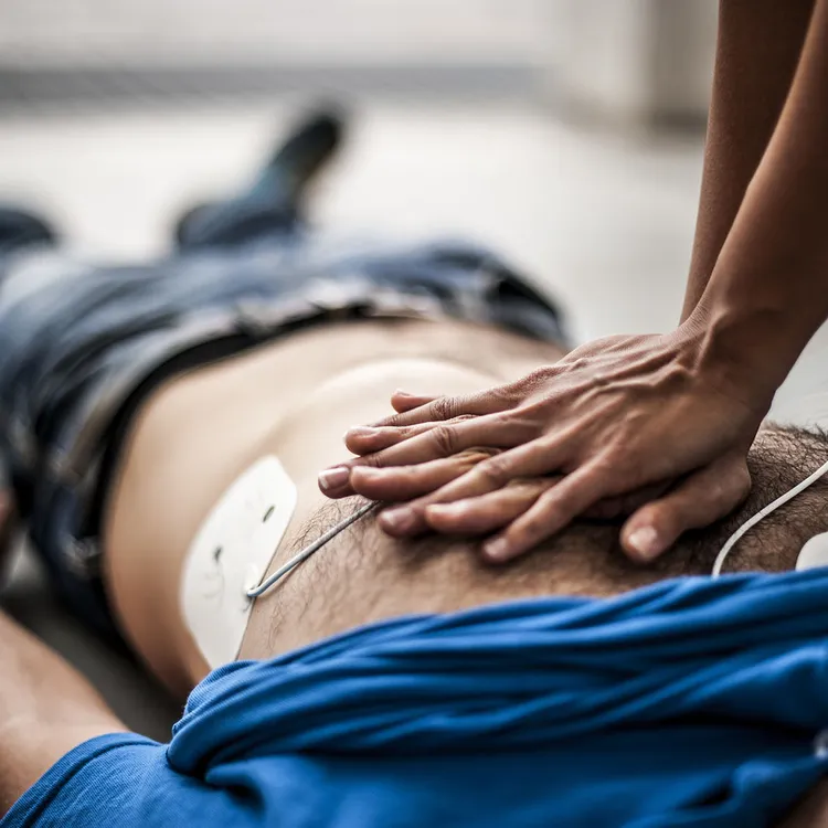 Eine Person zu sehen, die auf dem Boden liegt und Erste Hilfe erhält. Hände führen eine Herzmassage durch, während Elektroden auf der Brust angebracht sind.