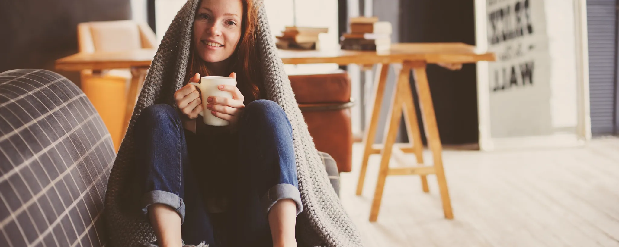 Eine Person sitzt gemütlich auf einem Sofa, in eine warme Decke gehüllt, und hält eine Tasse. Die herbstliche Atmosphäre lädt dazu ein, Hausmittel gegen Infekte zu genießen.