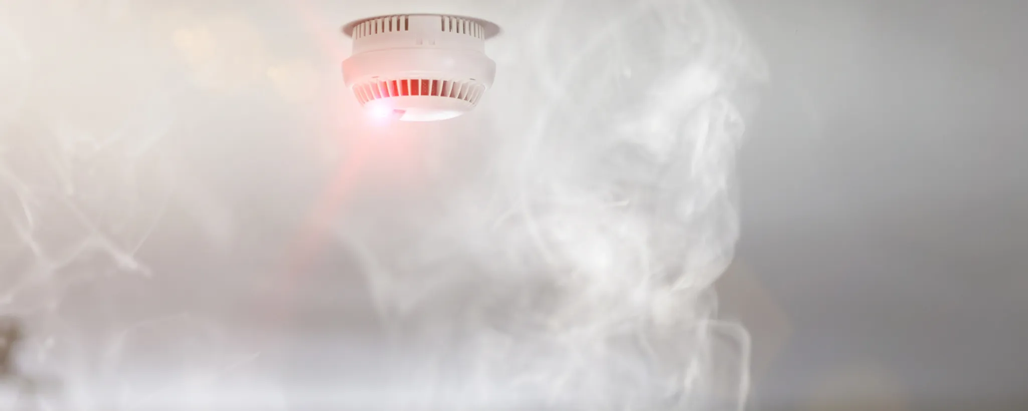 Einen aktivierten Rauchmelder mit rotem Licht, umgeben von Rauch. In Deutschland sind solche Geräte aufgrund gesetzlicher Vorschriften Pflicht, um die Sicherheit in Innenräumen zu gewährleisten.
