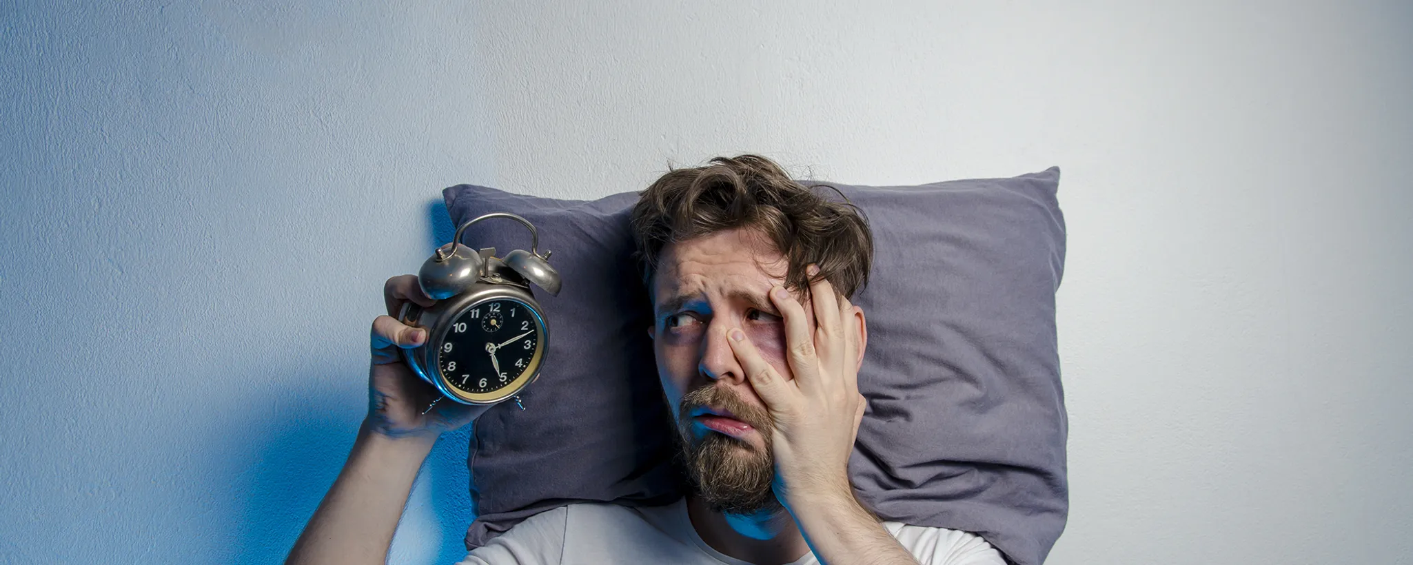 Ein Mann liegt im Bett und hält einen Wecker in der Hand, während er sich das Gesicht bedeckt. Die Szene deutet auf Schlafstörungen hin, da der Wecker eine späte Uhrzeit anzeigt.