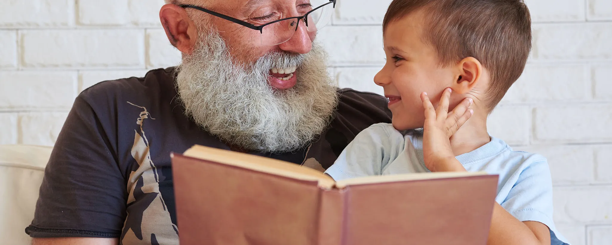 Ein älterer Mann liest einem Kind aus einem Buch vor, während sie auf einem Sofa sitzen. Die Szene vermittelt Geborgenheit und erinnert an den Wandel der Zeit, ähnlich wie Versicherungen Sicherheit bieten.
