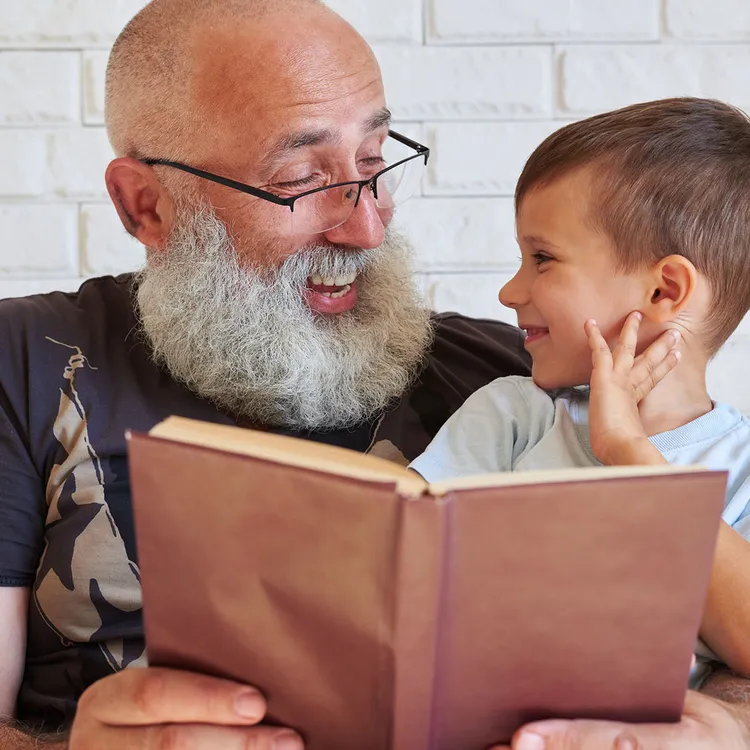 Ein älterer Mann liest einem Kind aus einem Buch vor, während sie auf einem Sofa sitzen. Die Szene vermittelt Geborgenheit und erinnert an den Wandel der Zeit, ähnlich wie Versicherungen Sicherheit bieten.
