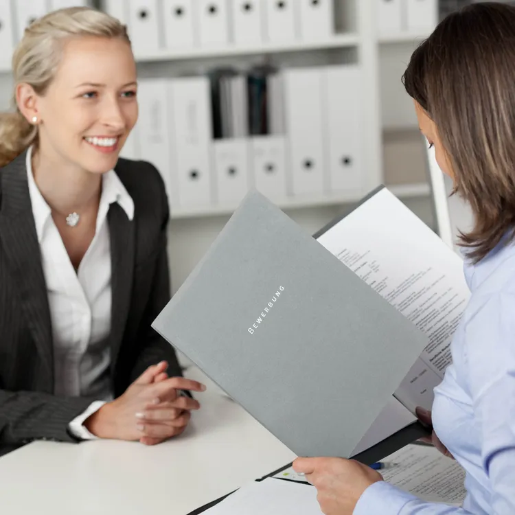 In einem Büro sitzt eine Person in einem grauen Anzug gegenüber einer anderen, die Bewerbungsunterlagen hält. Beide sind in ein Gespräch vertieft, während die Personalern die Unterlagen prüft. Im Hintergrund sind Aktenordner zu sehen.