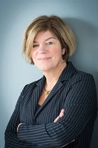 Die Leistungsfallbegleiterin Isabel Stabenow steht vor einer blaugrauen Wand, gekleidet in einem dunkel gestreiften Blazer und mit verschränkten Armen. Ihr blondes Haar fällt locker über die Schultern.