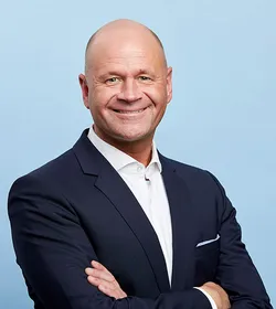 Oliver Blau, Gruppenleiter für Persönliche Beratung, steht mit verschränkten Armen in einem eleganten dunkelblauen Anzug vor einem hellblauen Hintergrund, wobei seine Rolle als Gruppenleiter für Persönliche Beratung betont wird.