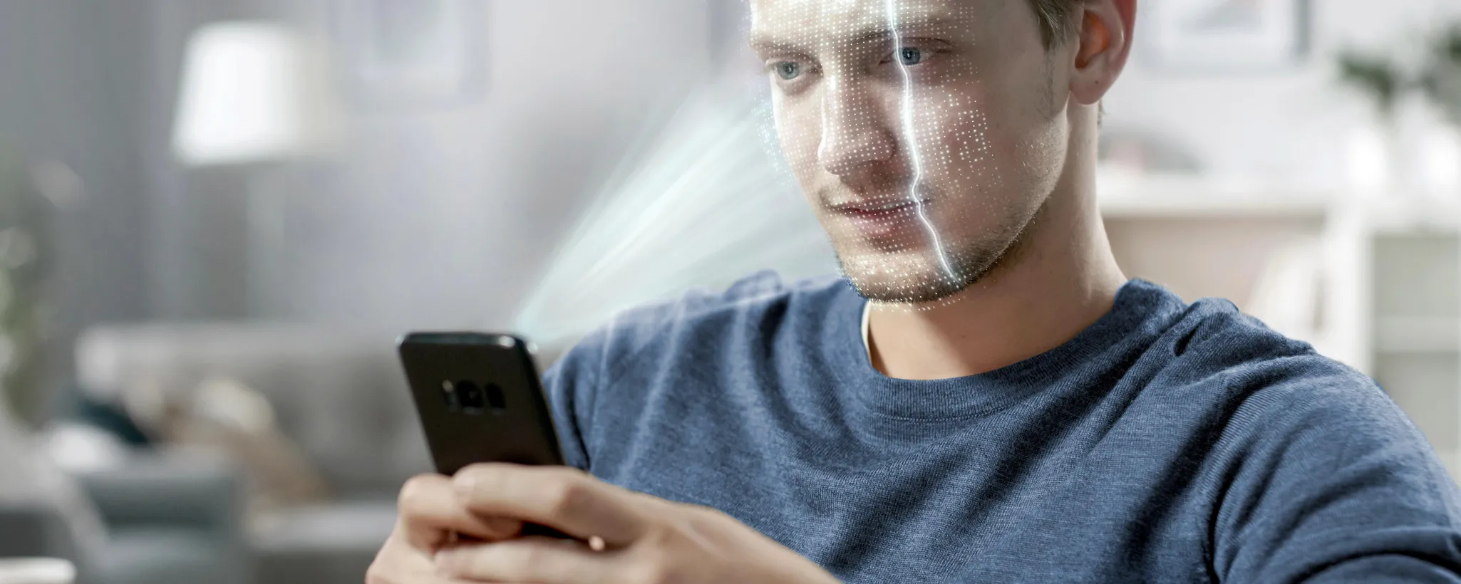 Ein Mann in einem blauen T-Shirt hält ein Smartphone, während er in einem gemütlichen Raum sitzt. Er nutzt die Safie-Funktion, um ein Selfie zu machen. Im Hintergrund sind ein Sofa und eine Lampe zu erkennen.