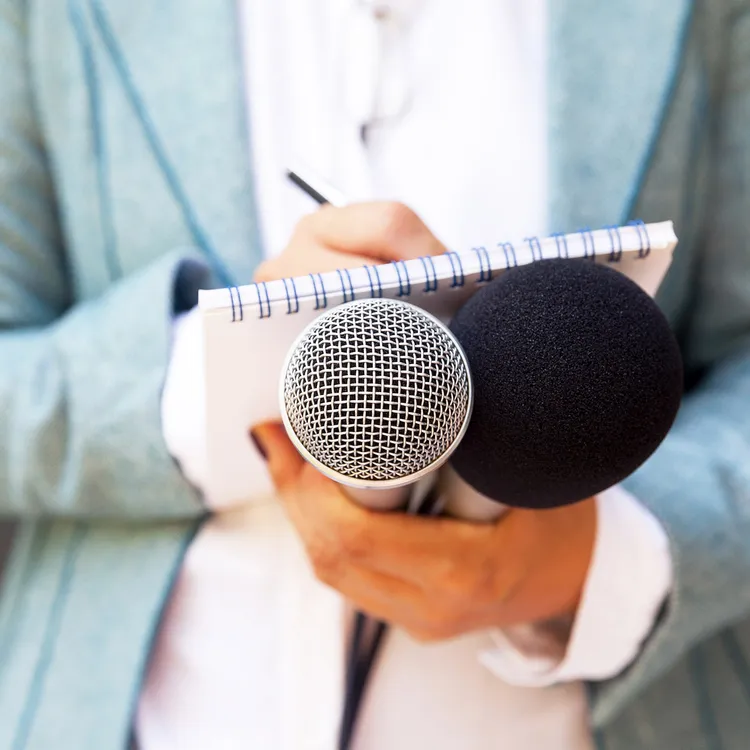 Eine Person in einem blauen Blazer hält zwei Mikrofone und ein Notizbuch, bereit für ein Interview, möglicherweise für eine Hannoversche Zeitung oder Medienplattform.