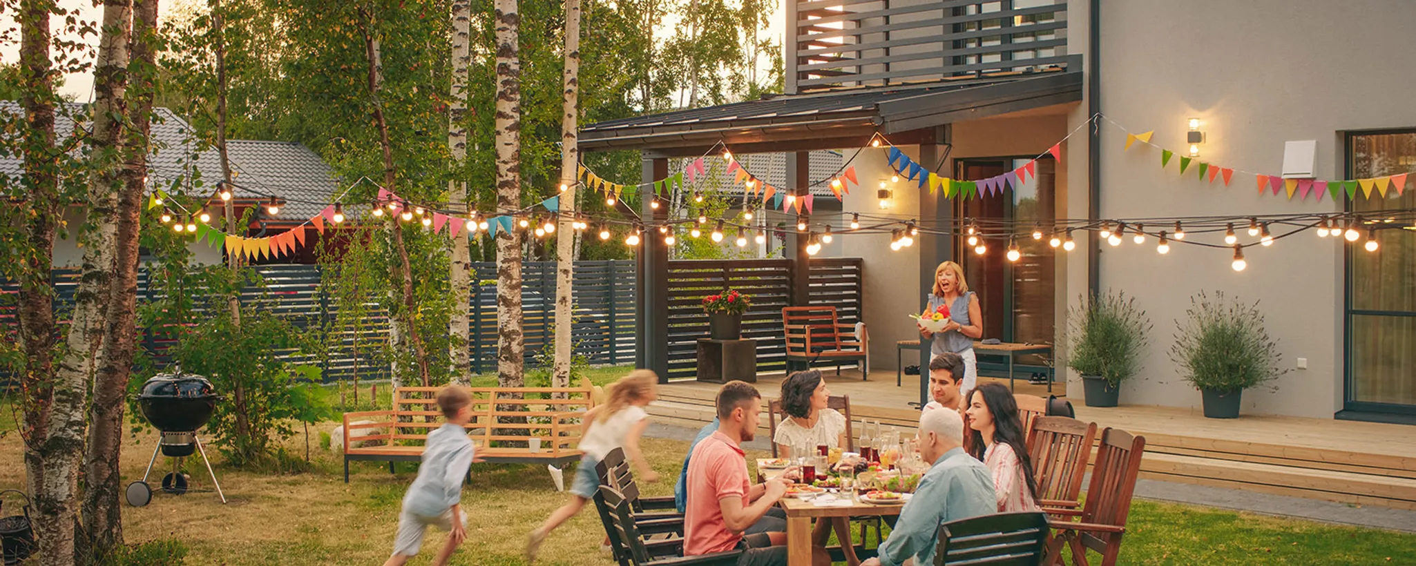 In einem modernen Eigenheim-Garten sitzt eine Familie unterschiedlichen Alters fröhlich um einen Tisch, während Kinder spielend vorbeilaufen. Bunte Lichterketten und Wimpel schmücken die Umgebung.