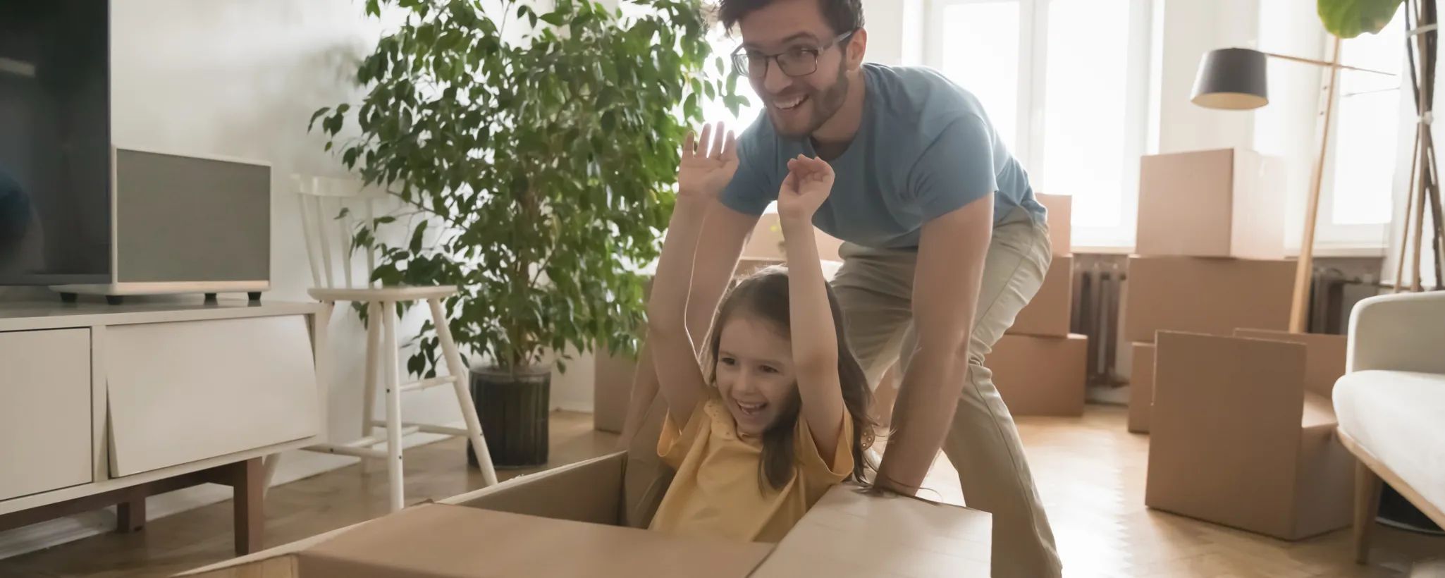 Ein Mann spielt fröhlich mit einem Kind, das in einem Umzugskarton sitzt. Auf der linken Seite steht in weißer Schrift auf hellblauem Hintergrund die Frage: „Wie kann man das Eigenheim absichern?“ Oben links ist ein Hashtag mit dem Label „Blogartikel“ zu sehen.